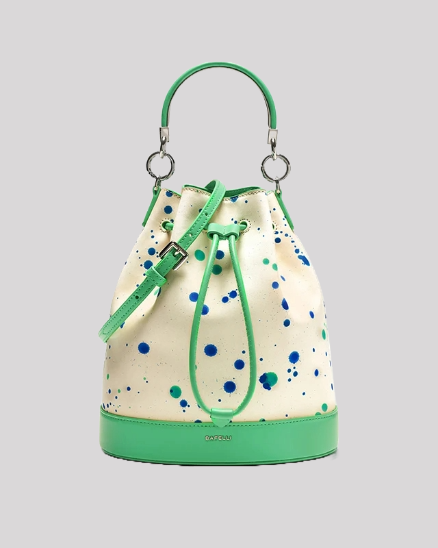 Front view of white bucket bag with green and blue paint splatter, green leather trim, silver handle rings, and drawstring closure by Bafelli