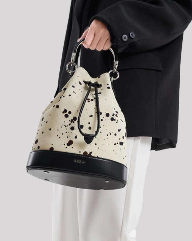 Model holding cream bucket bag by Bafelli with black paint splatter, black leather details and gold hardware top handle