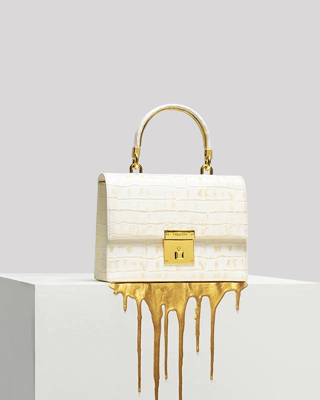 White croc-embossed top handle bag by Bafelli with gold lock shown on display pedestal with dripping gold paint detail