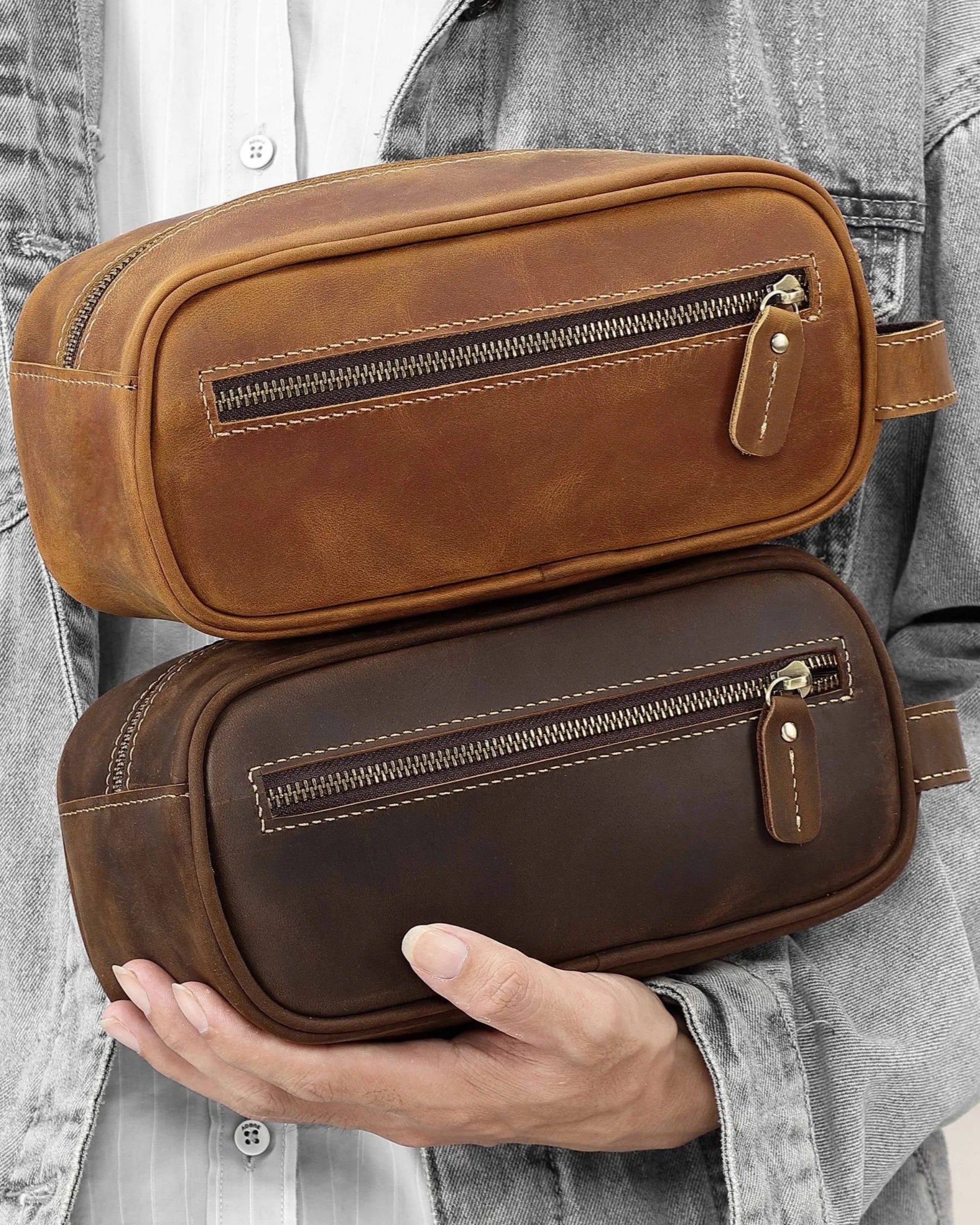 Person holding two leather toiletry bags in brown and dark brown, stacked for color comparison