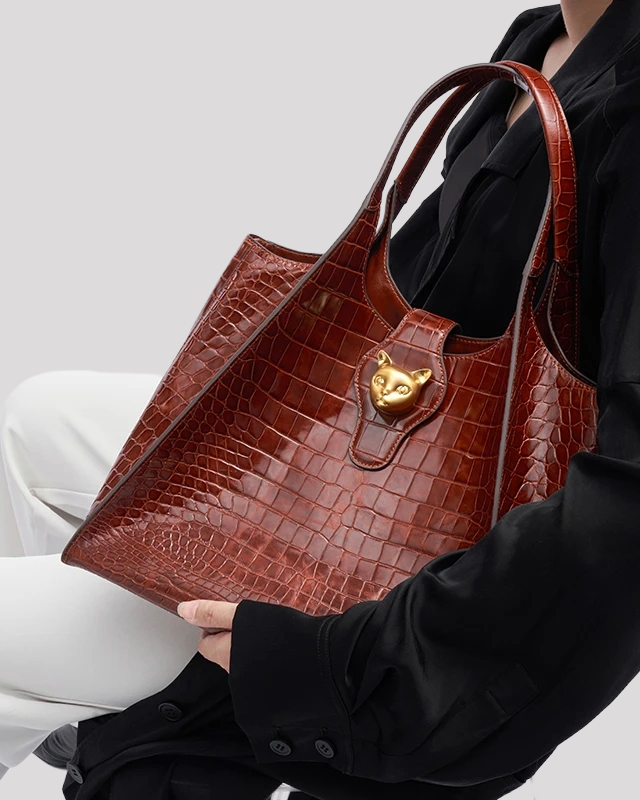 Brown croc-embossed tote bag by Bafelli with gold cat clasp, carried by model in black top and white pants