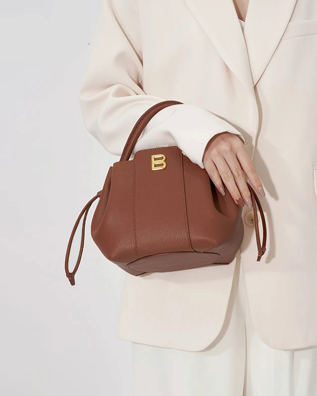 Model holding brown leather bucket bag with gold B logo, styled with cream blazer and white trousers