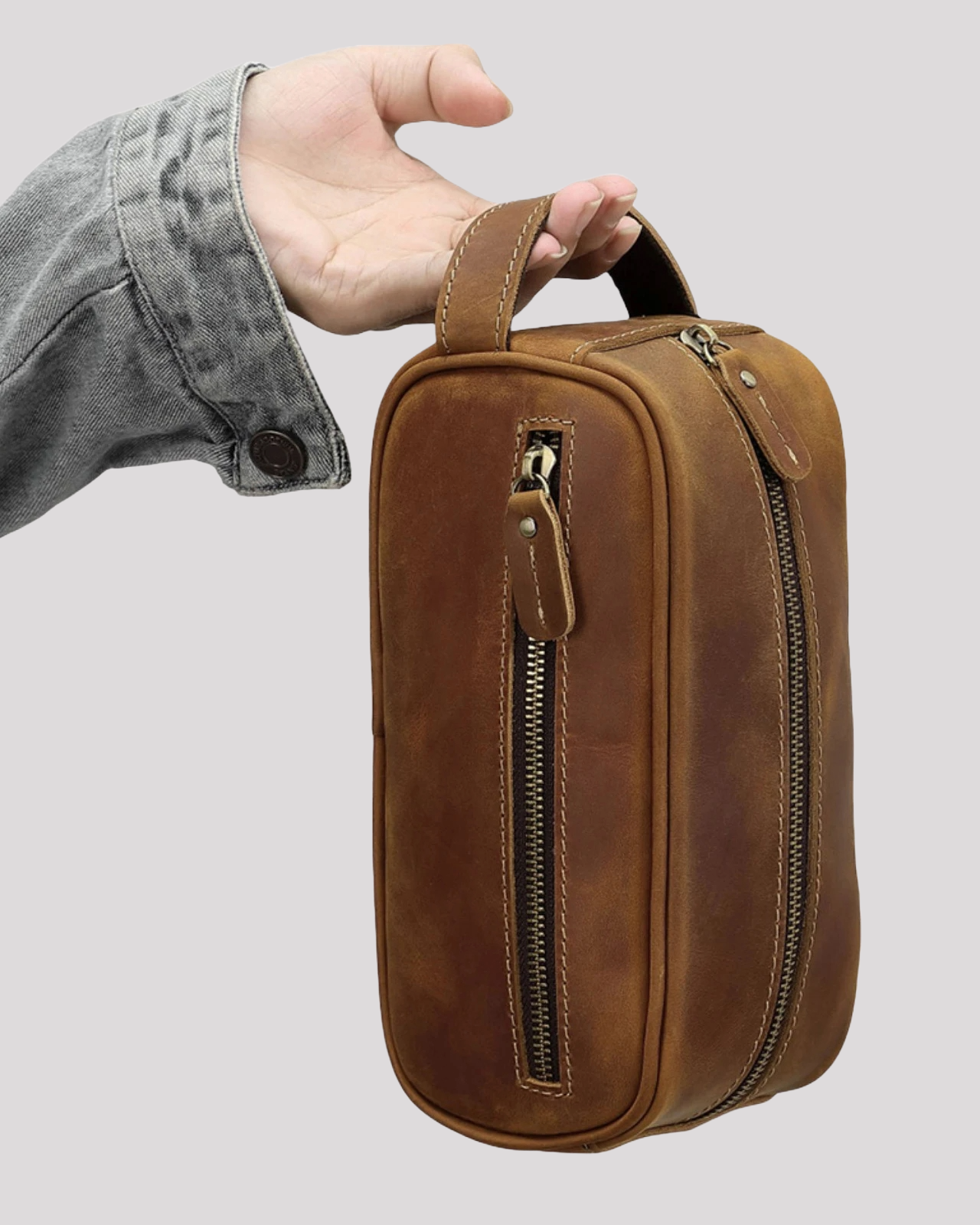Hand holding brown leather toiletry bag showing side zipper and compact shape