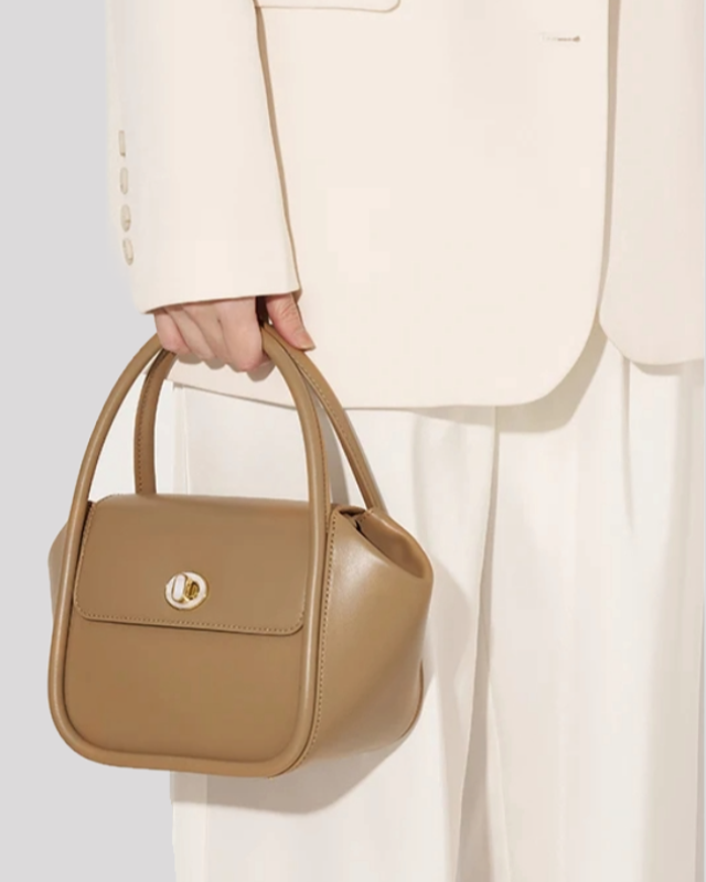 Woman holding taupe structured leather handbag by Bafelli with top handles, styled with a cream blazer and white pants