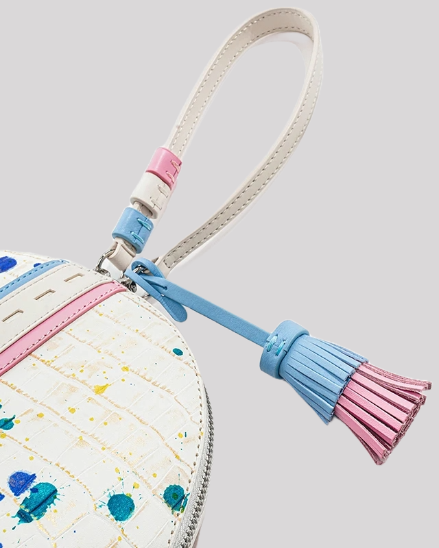 Close-up of blue and pink tassel detail on white crocodile-embossed wristlet bag by Bafelli with colorful paint splatter accents