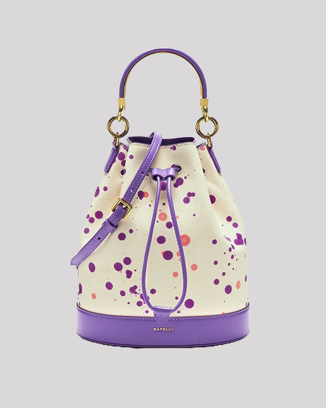Front view of white bucket bag by Bafelli with purple leather trim, paint-splatter design, gold handle rings, and adjustable strap